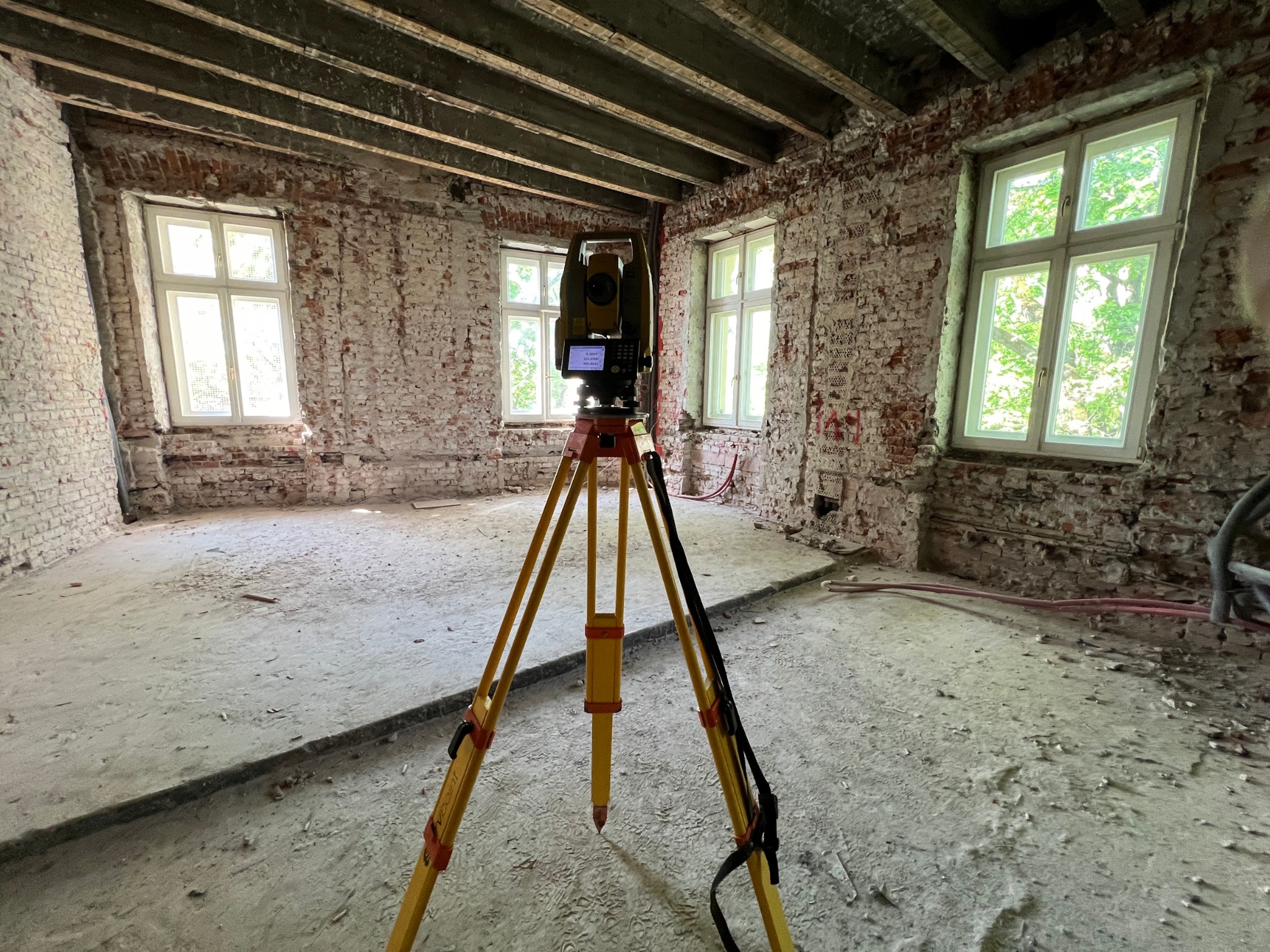 Żółty teodolit ustawiony na trójnogu wewnątrz budynku w trakcie remontu, widoczne surowe ceglane ściany z oknami i betonowa podłoga w stanie surowym.