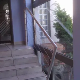 Schody wejściowe wyłożone płytkami z metalową balustradą z linkami stalowymi, widok z dołu na drzwi wejściowe i okno z widokiem na zieleń.