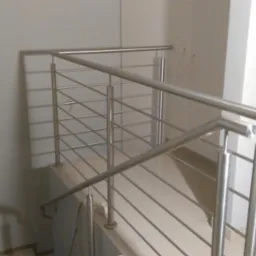 Nowoczesna balustrada schodowa ze stali nierdzewnej, z widocznymi spawami i mocowaniami, na tle jasnoszarej ściany i płytek podłogowych