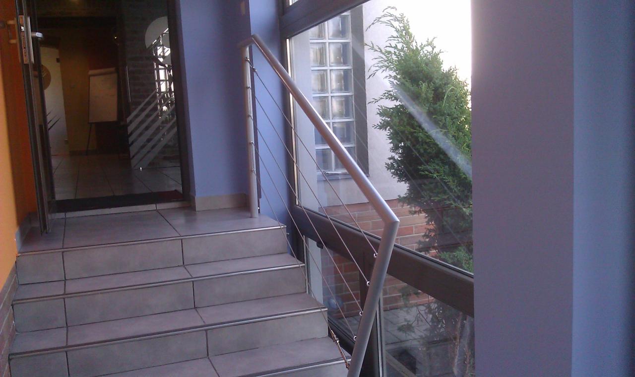 Schody wejściowe wyłożone płytkami z metalową balustradą z linkami stalowymi, widok z dołu na drzwi wejściowe i okno z widokiem na zieleń.