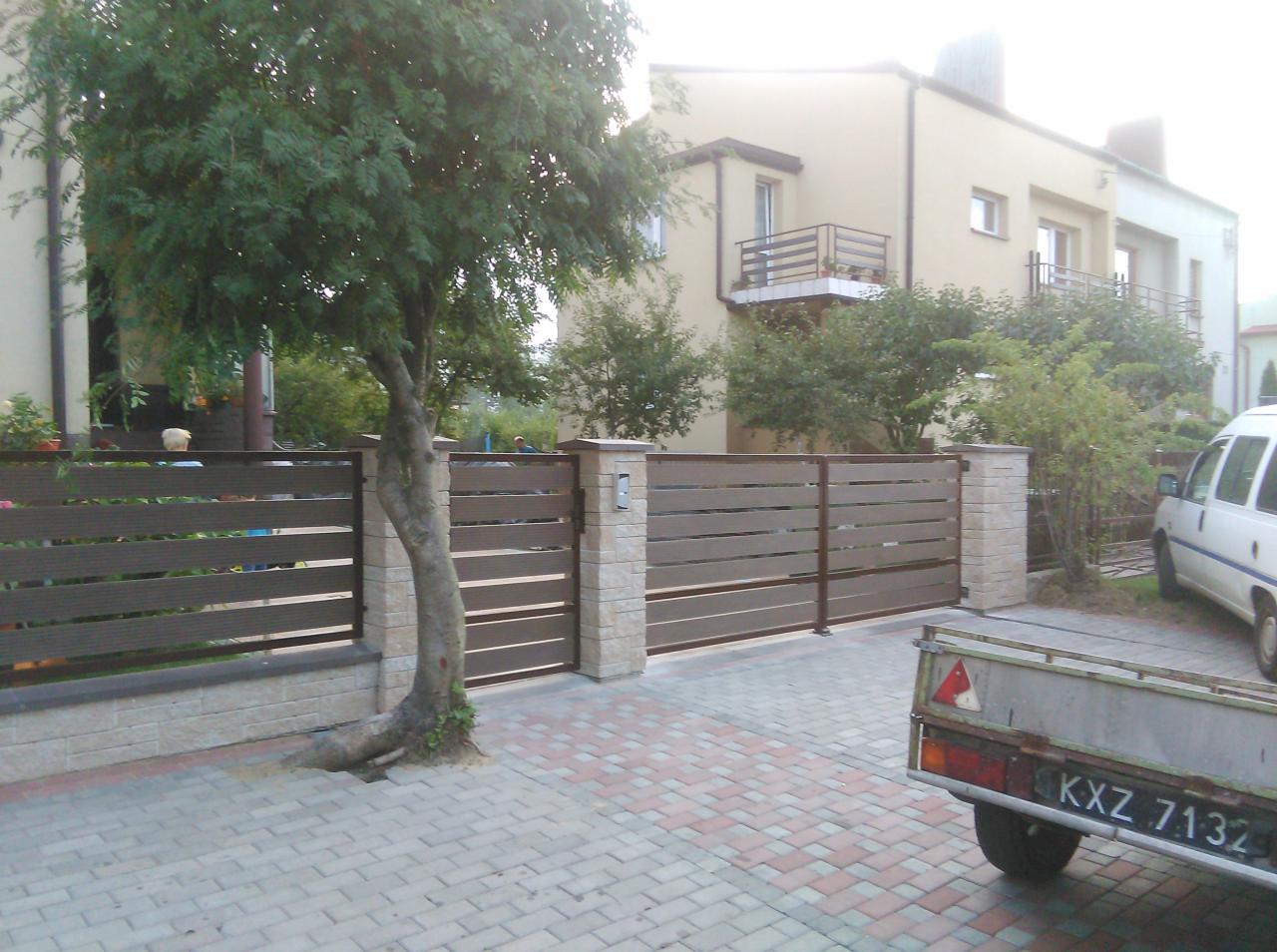 Nowoczesne ogrodzenie panelowe z poziomych desek kompozytowych w kolorze brązowym, osadzone na słupkach z imitacji kamienia, z drzewem rosnącym tuż przy jednym z przęseł, widok z zewnątrz posesji.