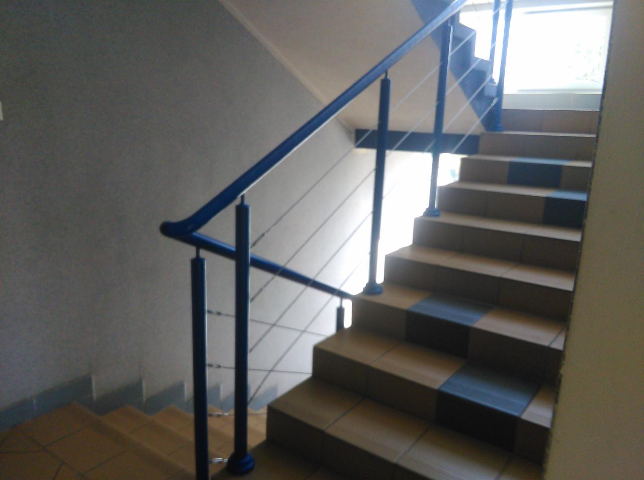 Klatka schodowa z ceramicznymi stopniami w odcieniach beżu i czerni, zabezpieczona niebieską, metalową balustradą z cienkimi, stalowymi linkami.