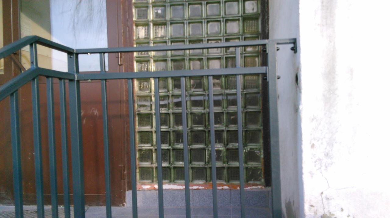 Zewnętrzna, metalowa balustrada schodowa w kolorze ciemnozielonym, z widoczną ścianą z luksferów w tle i fragmentem białej ściany z prawej strony.