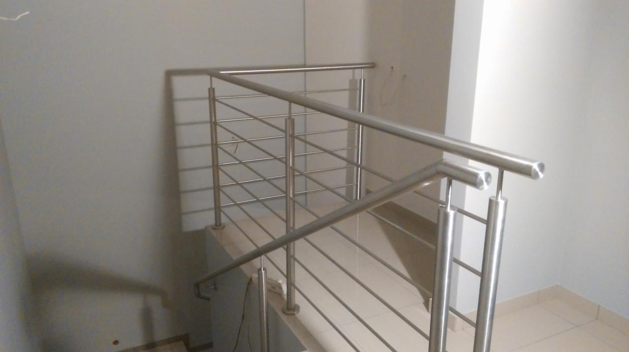 Nowoczesna balustrada schodowa ze stali nierdzewnej, z widocznymi spawami i mocowaniami, na tle jasnoszarej ściany i płytek podłogowych