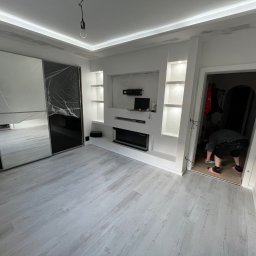 USŁUGI REMONTOWO-BUDOWLANE SEBASTIAN PIETKIEWICZ - Nowoczesny salon po remoncie: zabudowa TV z półkami, szafa z lustrami, jasne panele podłogowe. Wykończenie w bieli i czerni. Osoba w drzwiach.