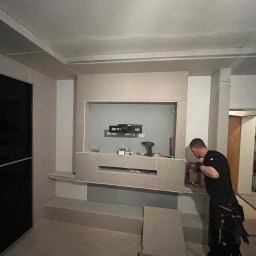 USŁUGI REMONTOWO-BUDOWLANE SEBASTIAN PIETKIEWICZ - Montaż płyty gipsowo-kartonowej na ścianie z wnęką na telewizor. Widoczny pracownik w trakcie pracy. W tle schody.