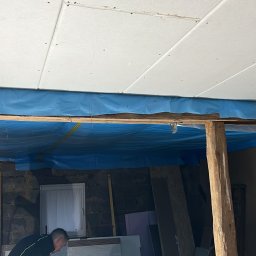 USŁUGI REMONTOWO-BUDOWLANE SEBASTIAN PIETKIEWICZ - Montaż płyt karton-gips na suficie, zabezpieczony folią. Widoczne drewniane belki i fragment pomieszczenia z oknem. Dwóch mężczyzn w trakcie pracy.