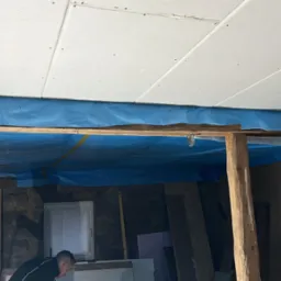 Montaż płyt karton-gips na suficie, zabezpieczony folią. Widoczne drewniane belki i fragment pomieszczenia z oknem. Dwóch mężczyzn w trakcie pracy.