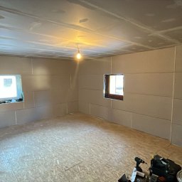 USŁUGI REMONTOWO-BUDOWLANE SEBASTIAN PIETKIEWICZ - Wnętrze pokoju z zamontowanymi płytami kartonowo-gipsowymi na ścianach i suficie, widoczne dwa okna i narzędzia na podłodze. Etap prac wykończeniowych.