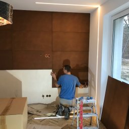 USŁUGI REMONTOWO-BUDOWLANE SEBASTIAN PIETKIEWICZ - Montaż paneli ściennych w brązowym odcieniu. Widoczny pracownik na kolanach, poziomica, drabina i niedokończona ściana. W tle okno z widokiem na zewnątrz.