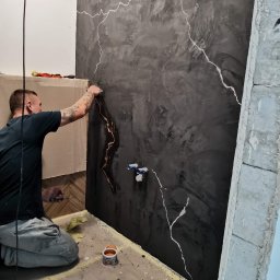 USŁUGI REMONTOWO-BUDOWLANE SEBASTIAN PIETKIEWICZ - Dekoracyjna ściana w łazience z efektem betonu i białymi żyłkami, wykonywana przez glazurnika. Widoczne narzędzia i fragment instalacji wodnej.