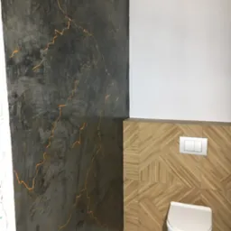 Wykończona ściana w łazience z dekoracyjnym tynkiem w odcieniach szarości i złotymi żyłkami, połączona z drewnopodobnymi płytkami i białą ceramiką.