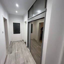 USŁUGI REMONTOWO-BUDOWLANE SEBASTIAN PIETKIEWICZ - Jasny korytarz po remoncie z szarą podłogą imitującą drewno i szafą wnękową z lustrzanymi drzwiami przesuwnymi. Widoczne drzwi wejściowe i oświetlenie sufitowe.