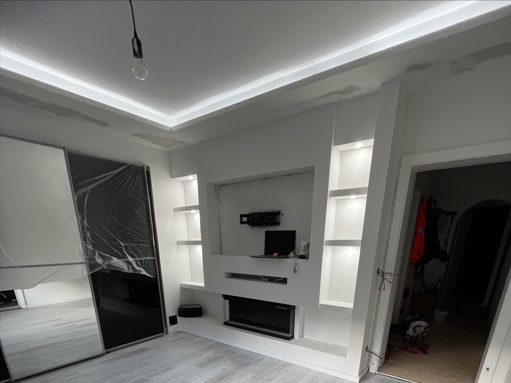 Nowoczesny salon po remoncie: białe ściany, zabudowa TV z półkami, oświetlenie LED i lustrzana szafa przesuwna. Widoczne niedoskonałości wykończenia.