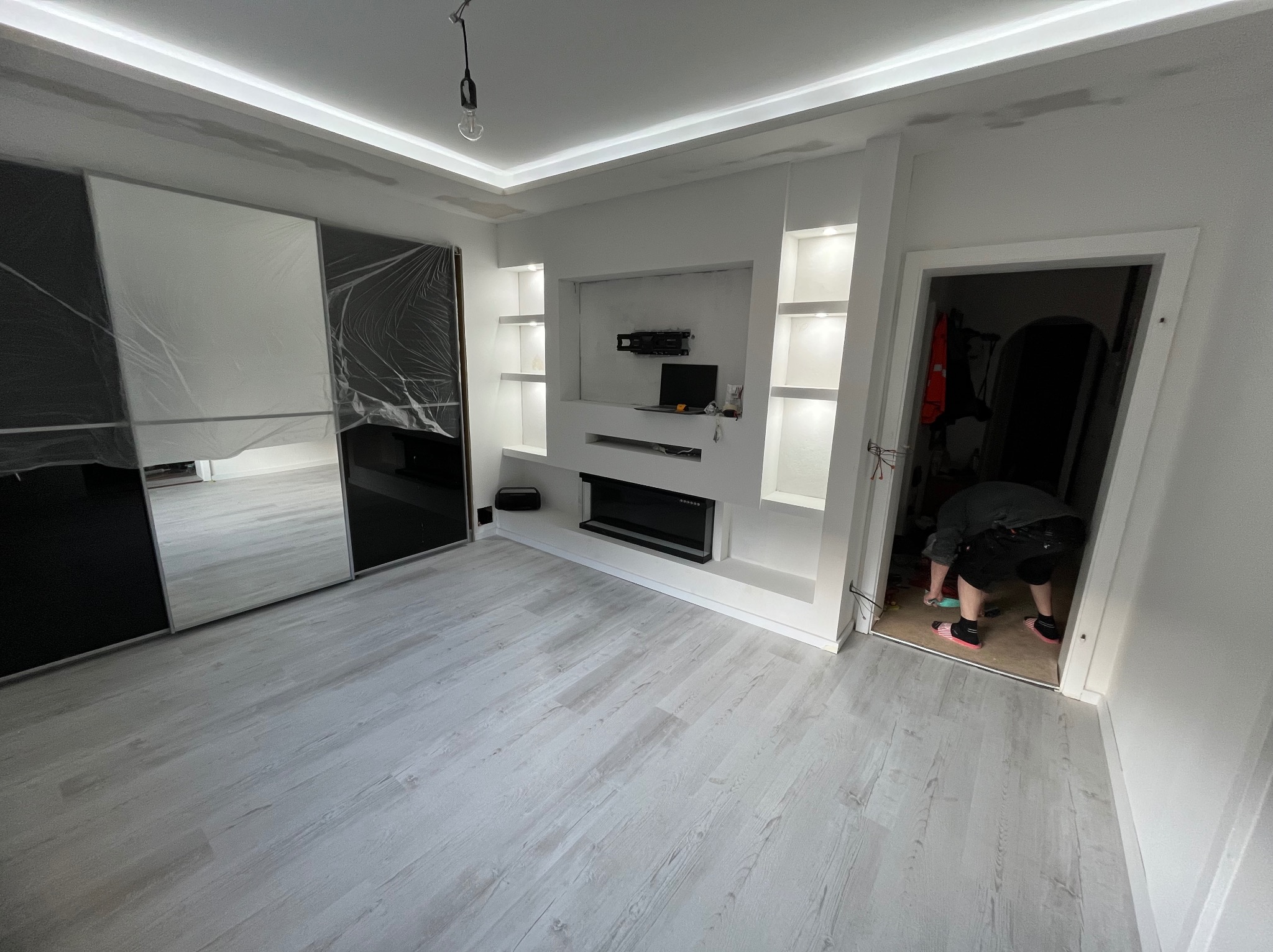 Nowoczesny salon po remoncie: zabudowa TV z półkami, szafa z lustrami, jasne panele podłogowe. Wykończenie w bieli i czerni. Osoba w drzwiach.