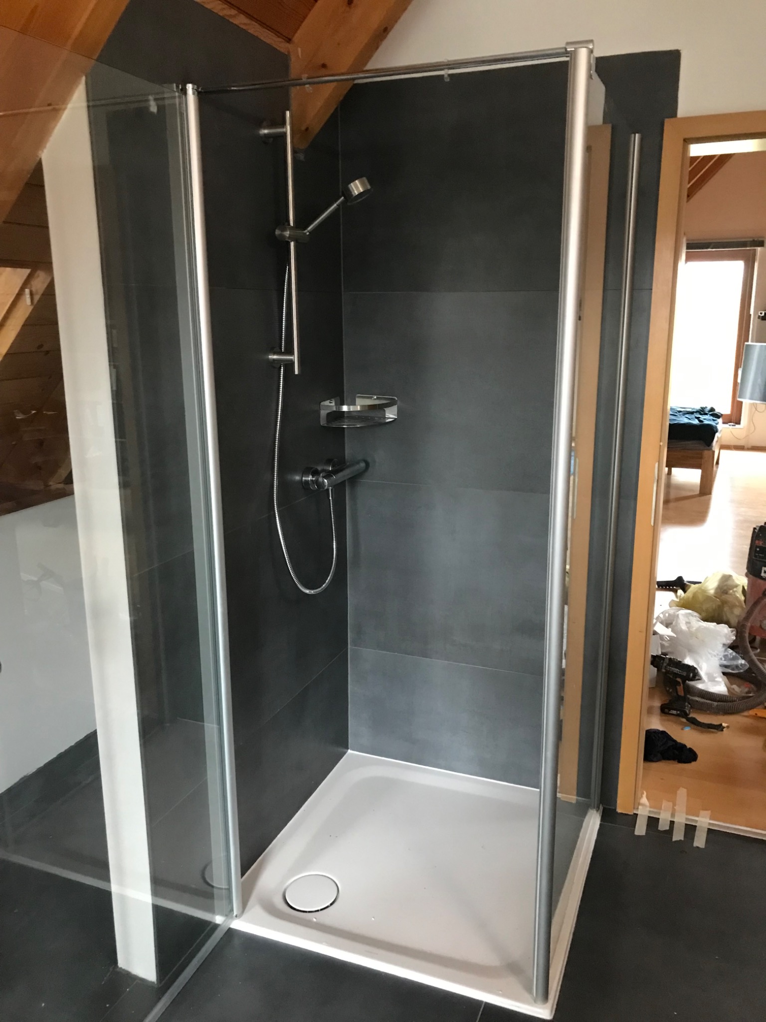 Kabina prysznicowa z szarymi płytkami w łazience na poddaszu. Widoczny brodzik, bateria i deszczownica. W tle otwarte drzwi do pokoju.