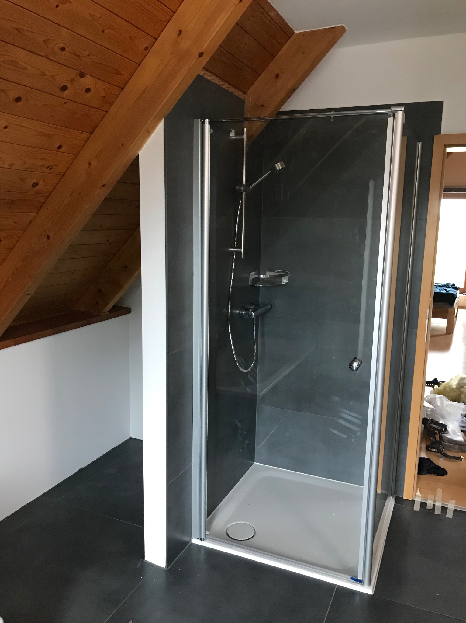 Kabina prysznicowa z szarymi płytkami w łazience na poddaszu, widoczna drewniana więźba dachowa. Ukończony remont z nowoczesnym designem.