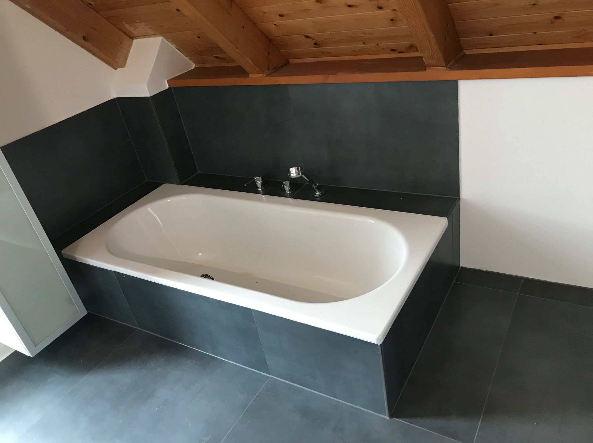 Wykończona łazienka na poddaszu: biała wanna obudowana ciemnymi płytkami, szare płytki na podłodze, drewniany sufit.