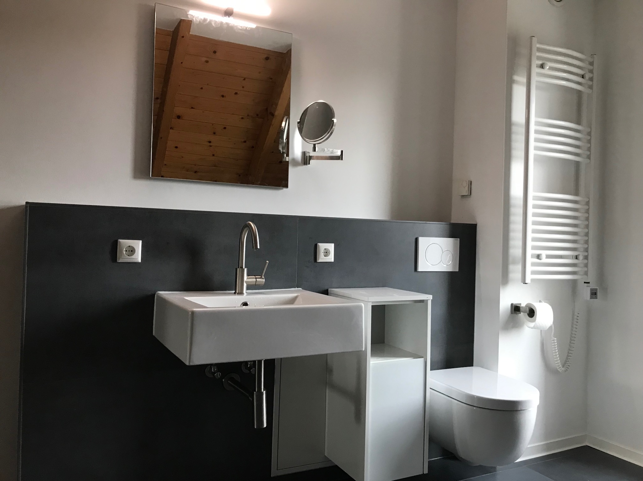 Nowoczesna łazienka z białą umywalką, toaletą i grzejnikiem drabinkowym. W lustrze odbija się drewniany strop. Szara ściana za umywalką, minimalistyczny design.