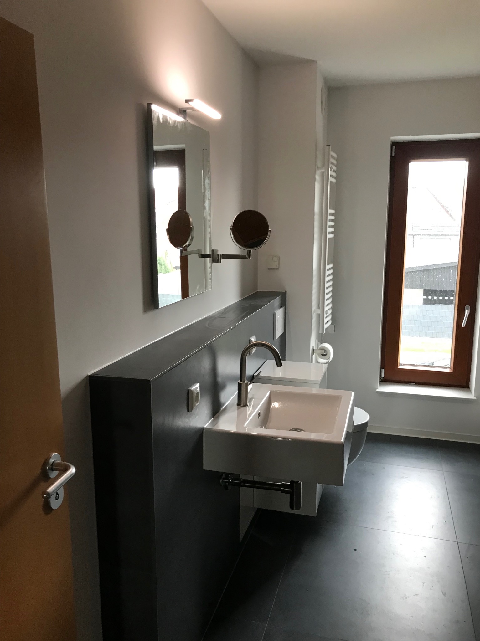 Nowoczesna łazienka z kwadratową umywalką, lustrem z oświetleniem i grzejnikiem drabinkowym. Ciemna zabudowa umywalki i podłoga kontrastują z jasnymi ścianami.