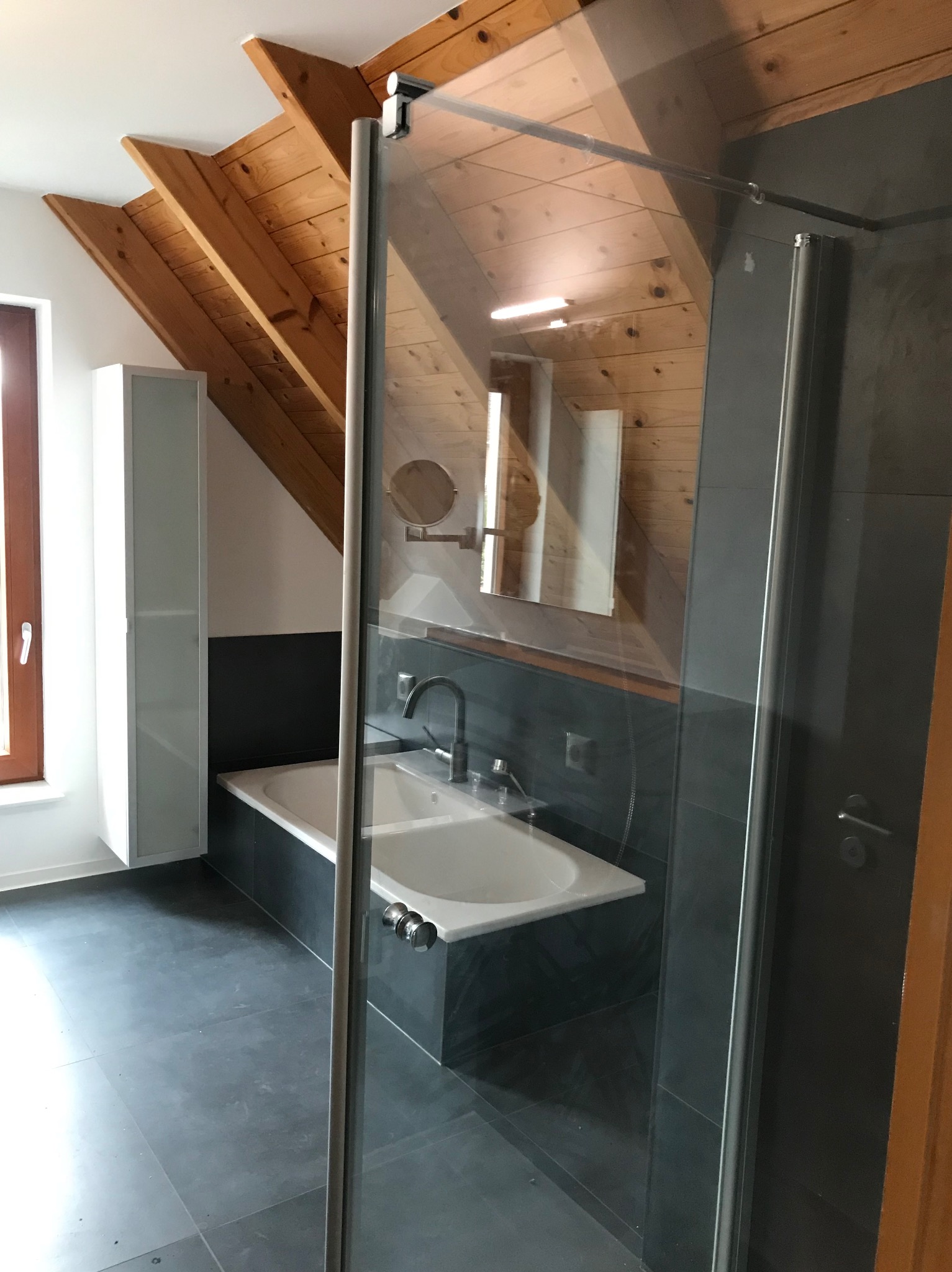 Nowoczesna łazienka na poddaszu z kabiną prysznicową, białą wanną i drewnianym wykończeniem skosu dachu. Szare płytki podłogowe i ciemne ściany.