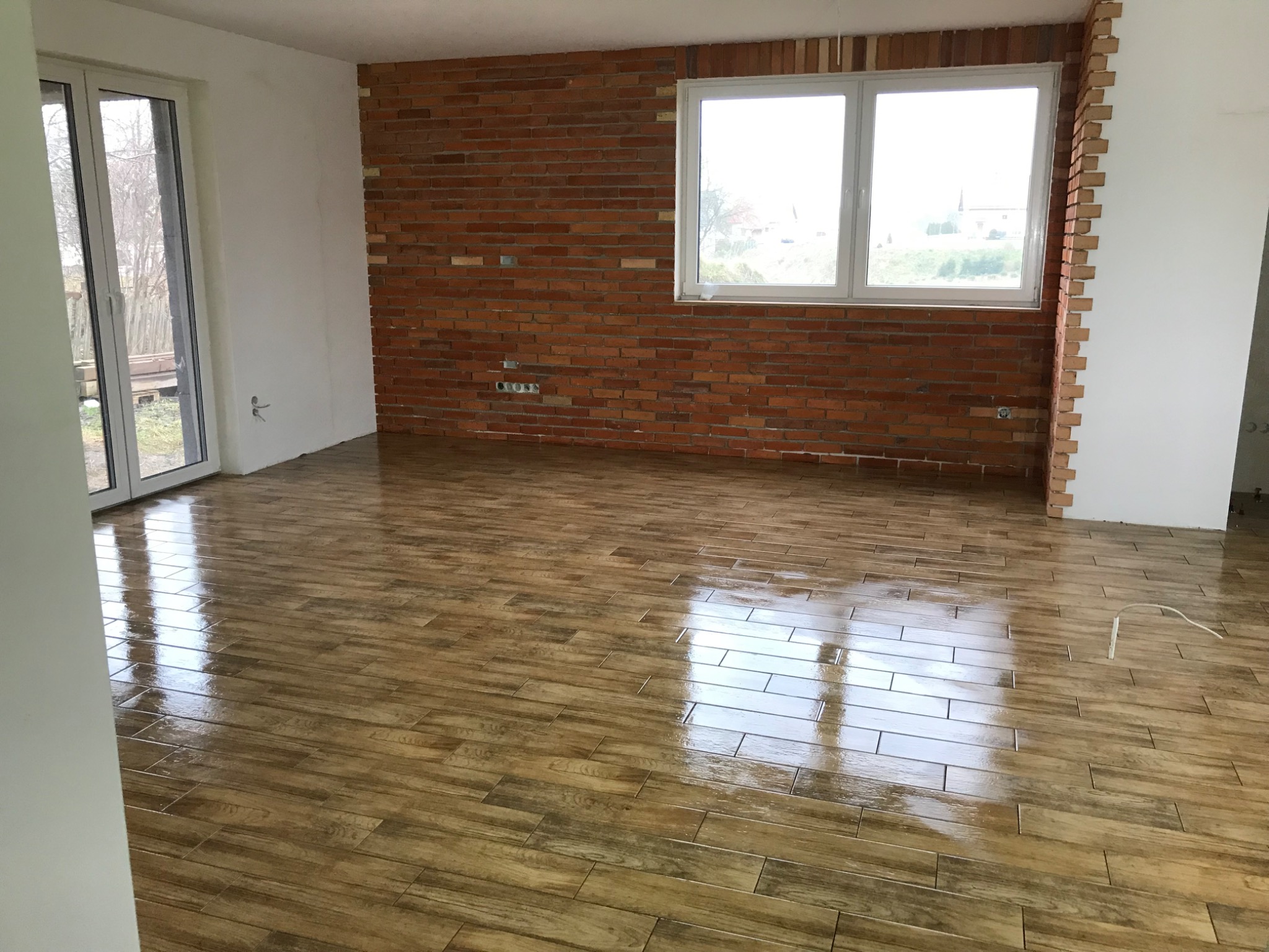 Pokój po remoncie: błyszcząca podłoga z paneli imitujących drewno, ściana z cegły i białe ściany. Widoczne okno i drzwi balkonowe.