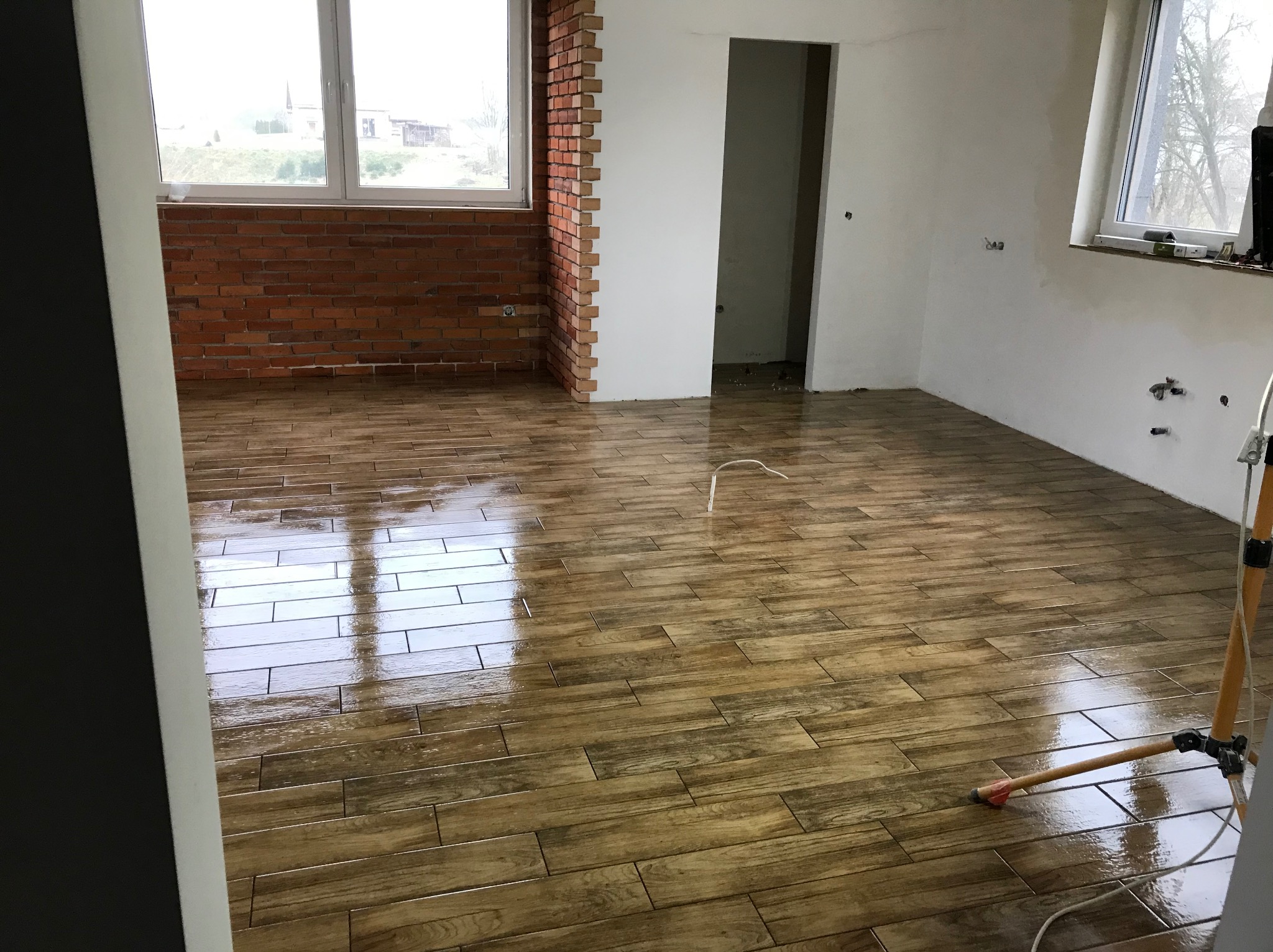 Ułożona podłoga z drewnopodobnych płytek w jasnym pokoju, fragment ściany z cegły, okno z widokiem na zewnątrz. Widoczny statyw oświetleniowy.