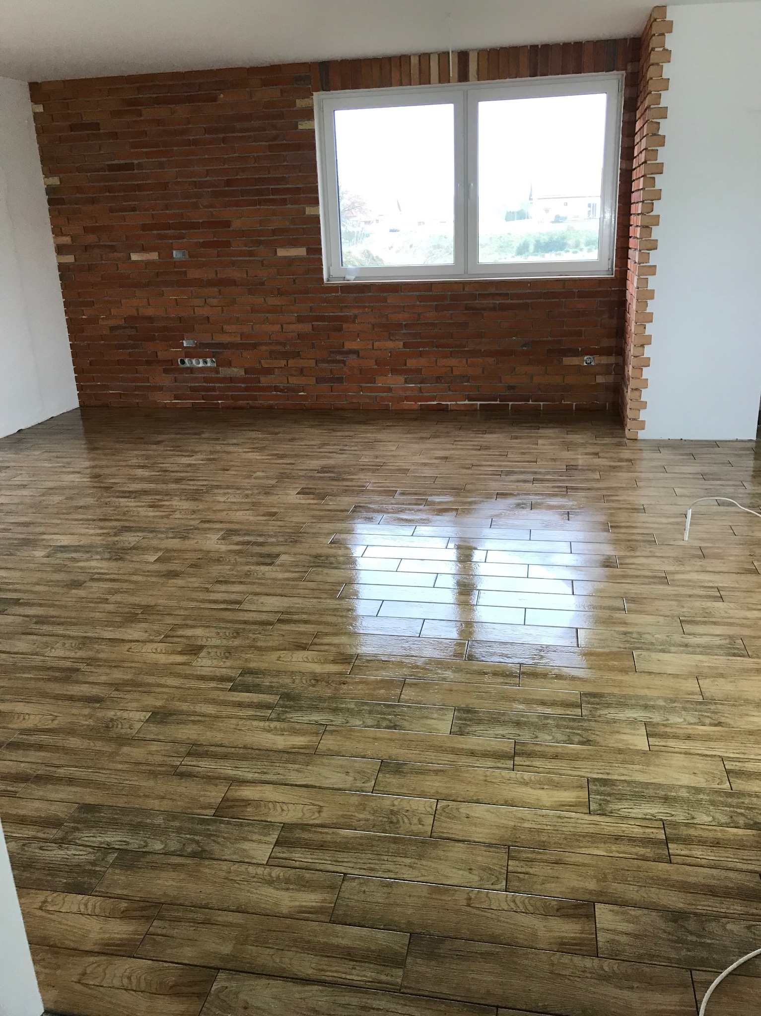 Połyskująca, drewnopodobna podłoga z płytek, ceglana ściana z oknem. Widoczne gniazdka elektryczne. Wykończone wnętrze w surowym stylu.