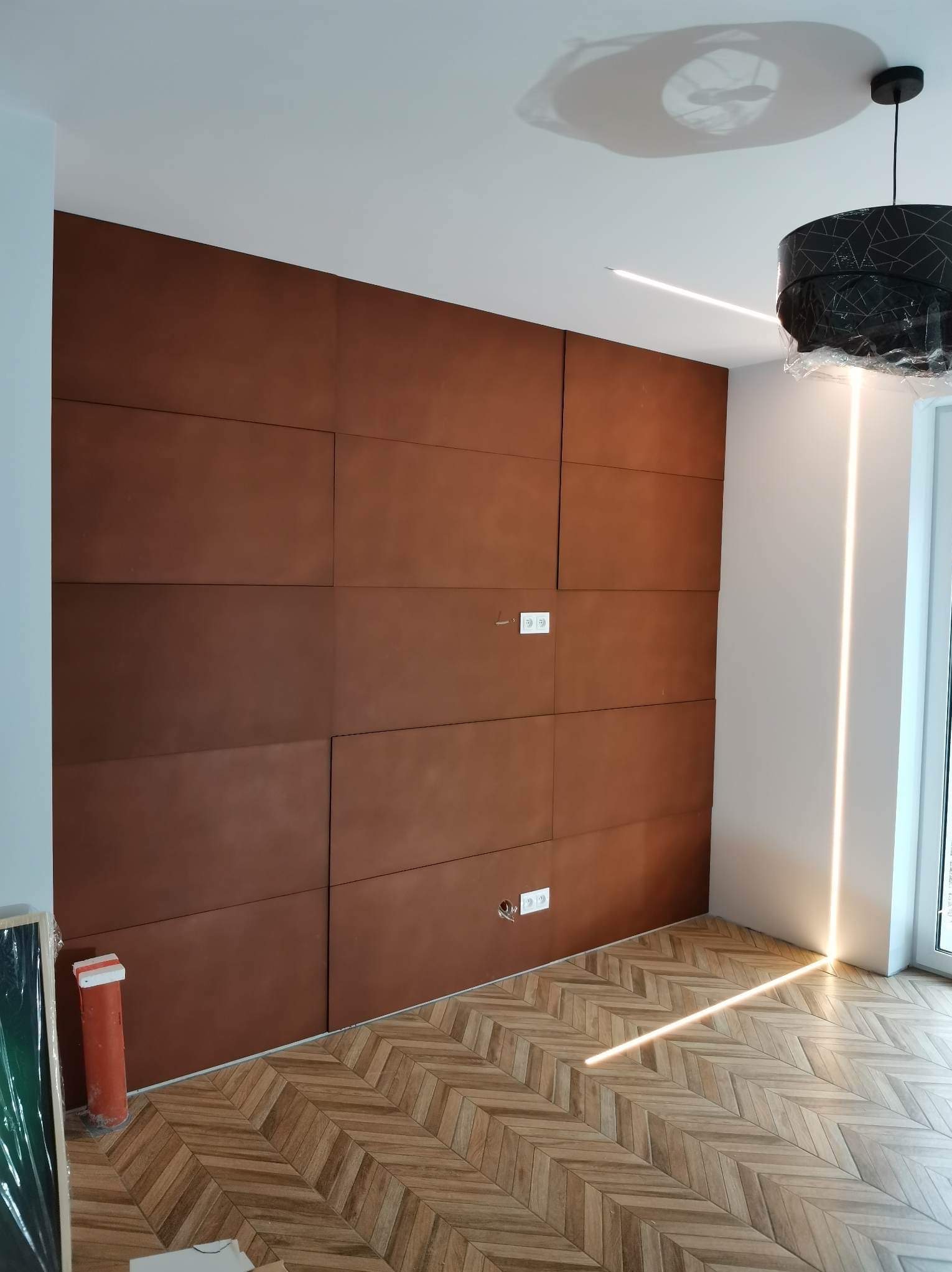 Panelowa ściana w odcieniach brązu i parkiet ułożony w jodełkę, tworzące nowoczesne wnętrze w Lęborku. Widoczne gniazdka elektryczne i oświetlenie LED.