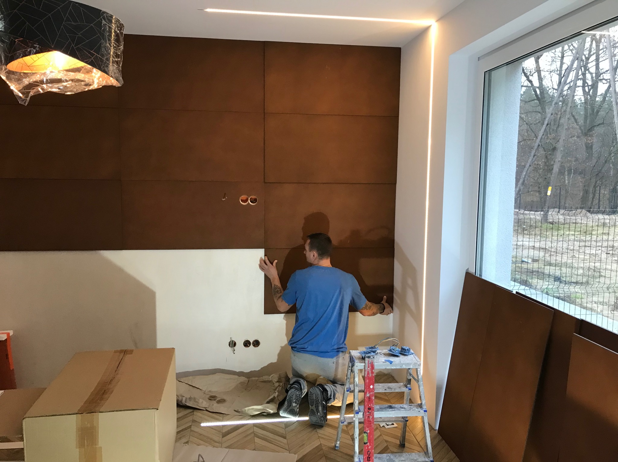 Montaż paneli ściennych w brązowym odcieniu. Widoczny pracownik na kolanach, poziomica, drabina i niedokończona ściana. W tle okno z widokiem na zewnątrz.