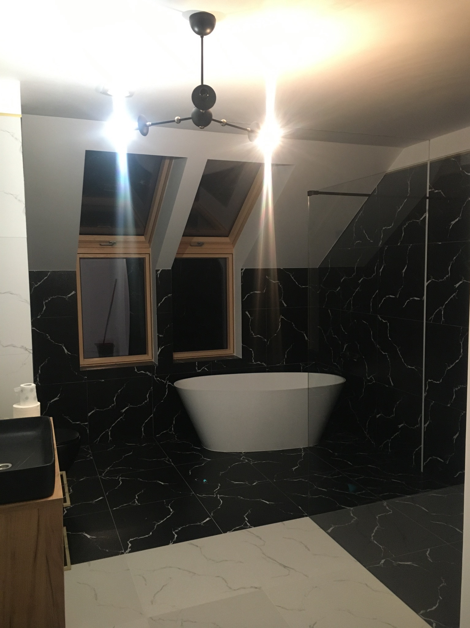 Nowoczesna łazienka na poddaszu z czarno-białymi płytkami imitującymi marmur, wolnostojącą wanną i oknami dachowymi. Minimalistyczne oświetlenie.