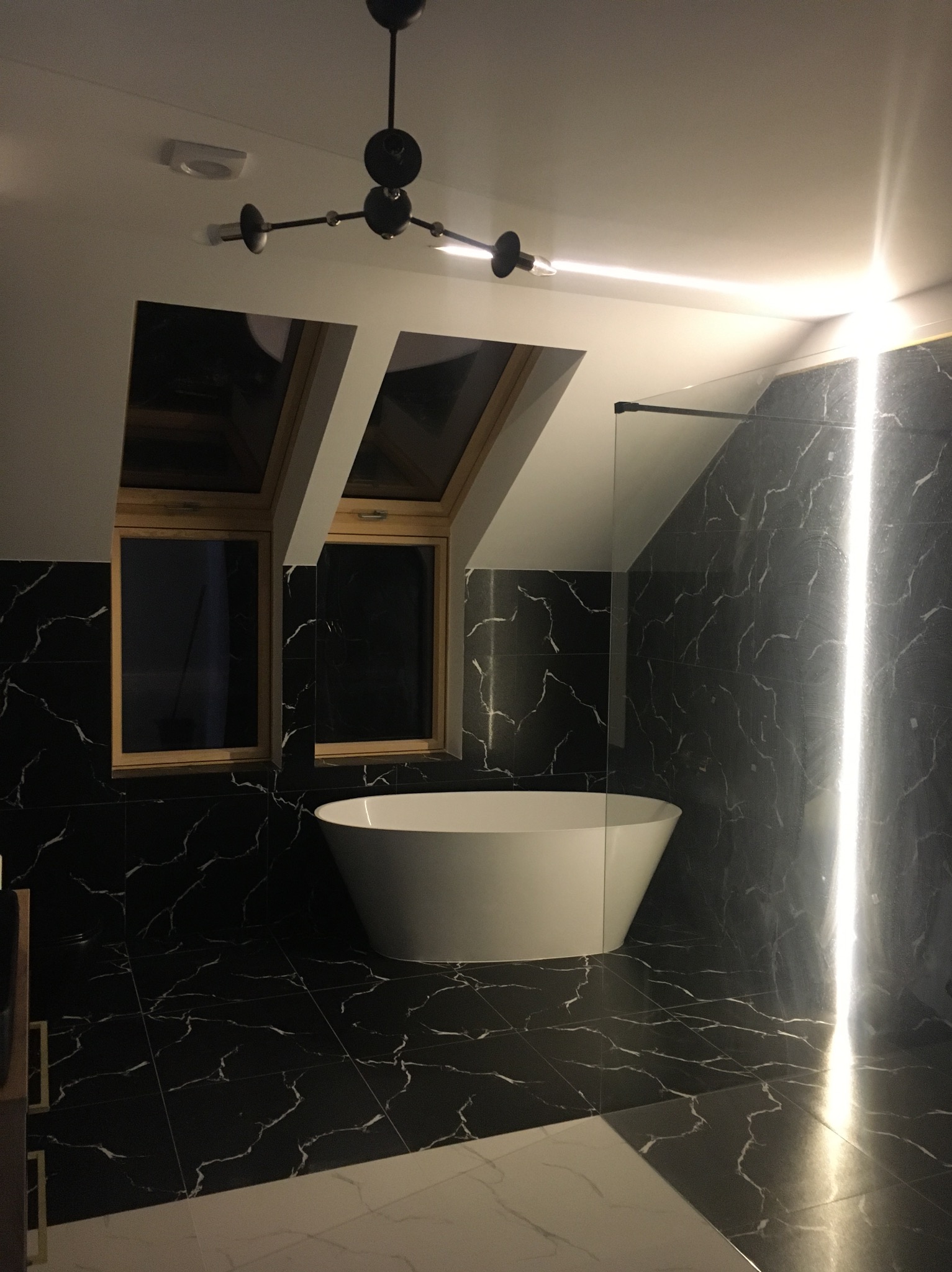 Elegancka łazienka na poddaszu z czarnymi płytkami imitującymi marmur, wolnostojącą białą wanną i nowoczesnym oświetleniem. Dwa okna dachowe w drewnianych ramach.