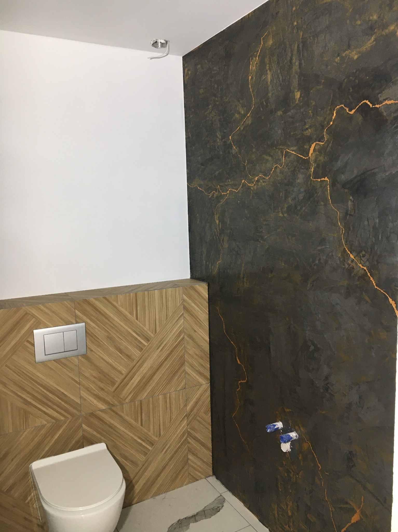 Nowoczesna łazienka z dekoracyjną, ciemną ścianą z efektem żyłkowania, drewnopodobnymi płytkami i podwieszaną toaletą. Widoczne przyłącza wodne.