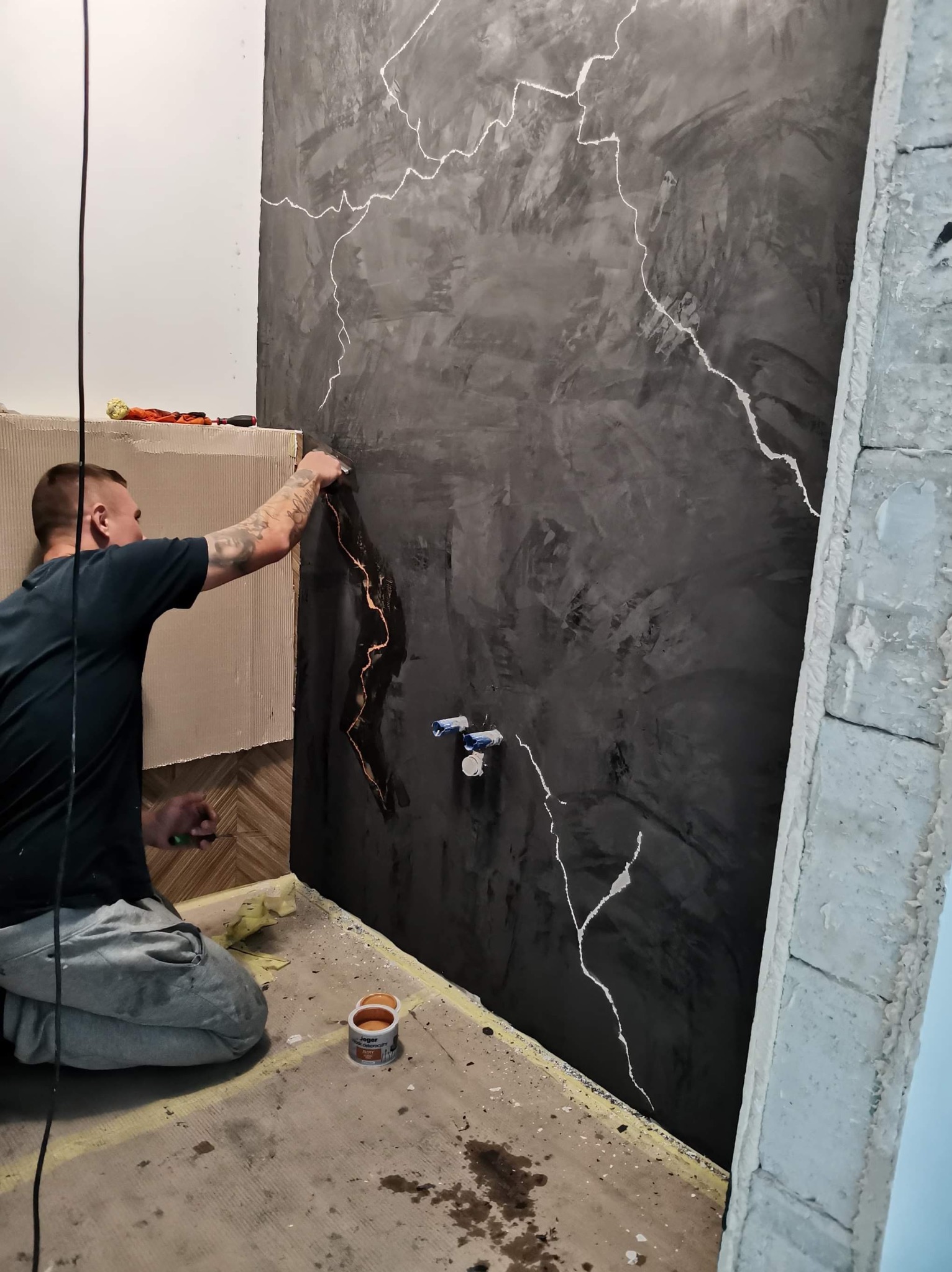Dekoracyjna ściana w łazience z efektem betonu i białymi żyłkami, wykonywana przez glazurnika. Widoczne narzędzia i fragment instalacji wodnej.