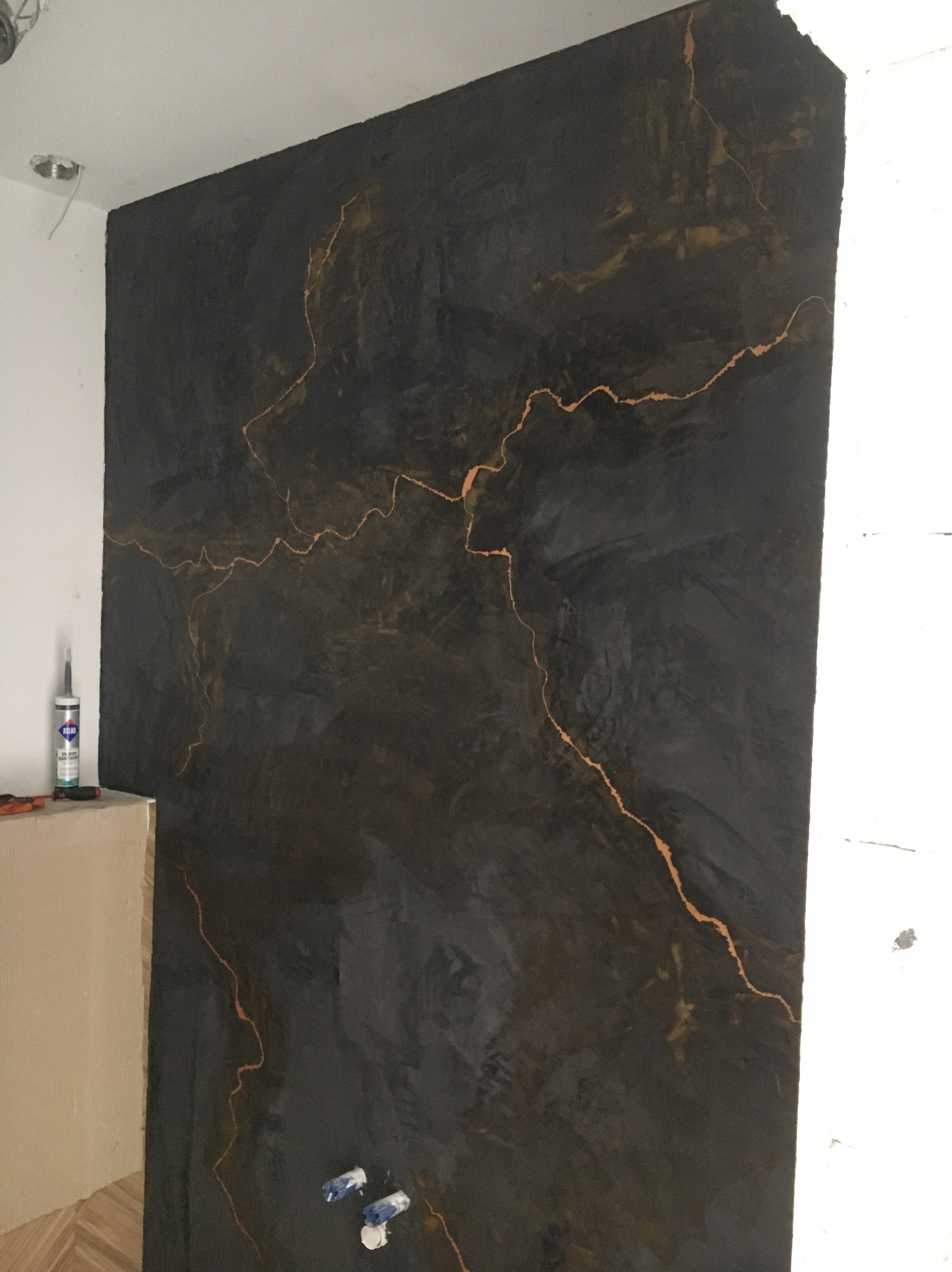 Czarna ściana z efektem żyłkowanego kamienia w odcieniach złota, wykonana techniką dekoracyjną. W tle widoczne narzędzia i materiały budowlane.