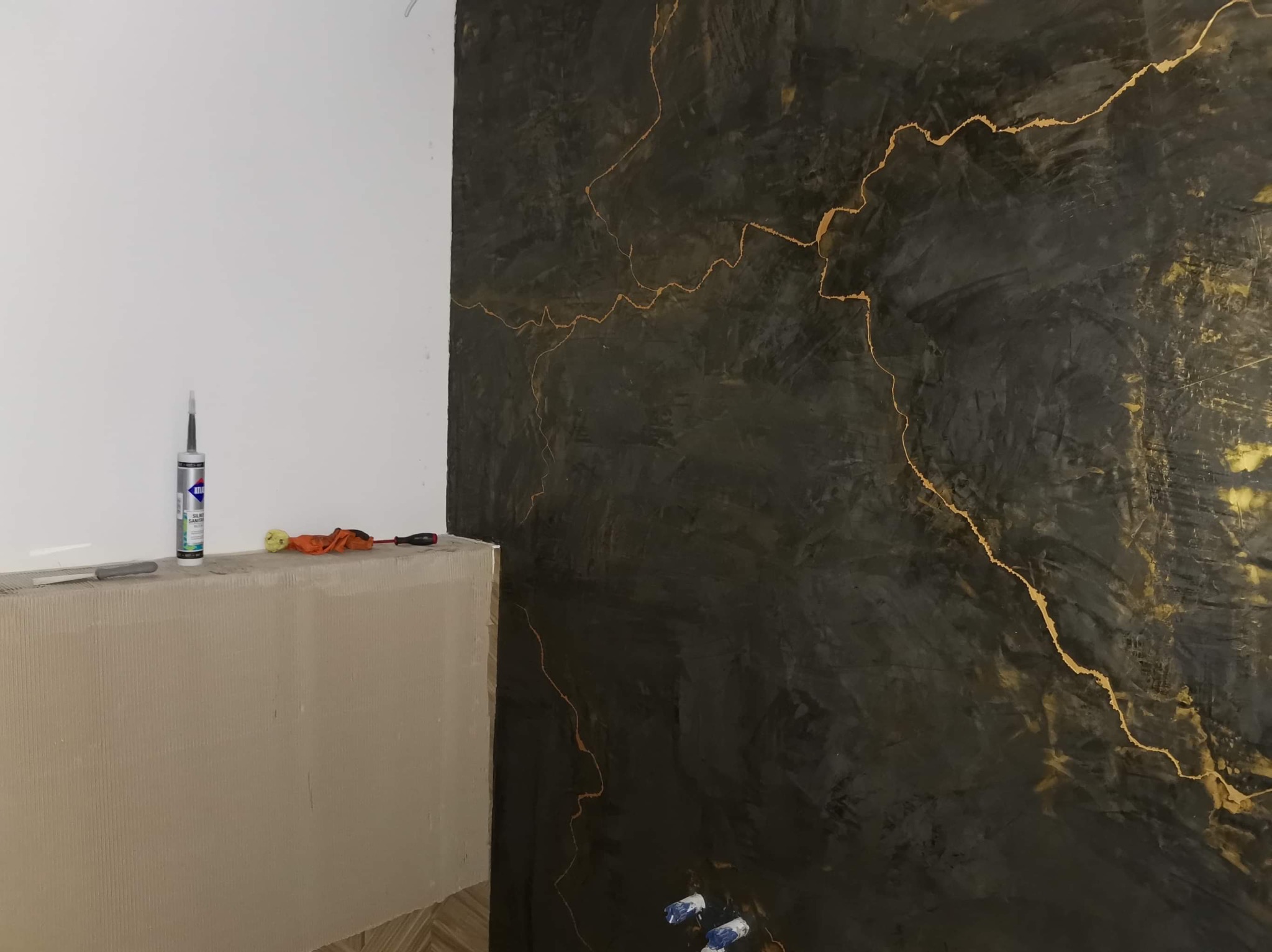 Część ściany z dekoracyjną czarną farbą imitującą marmur ze złotymi żyłkami oraz fragment przygotowanej pod płytki ściany z narzędziami.