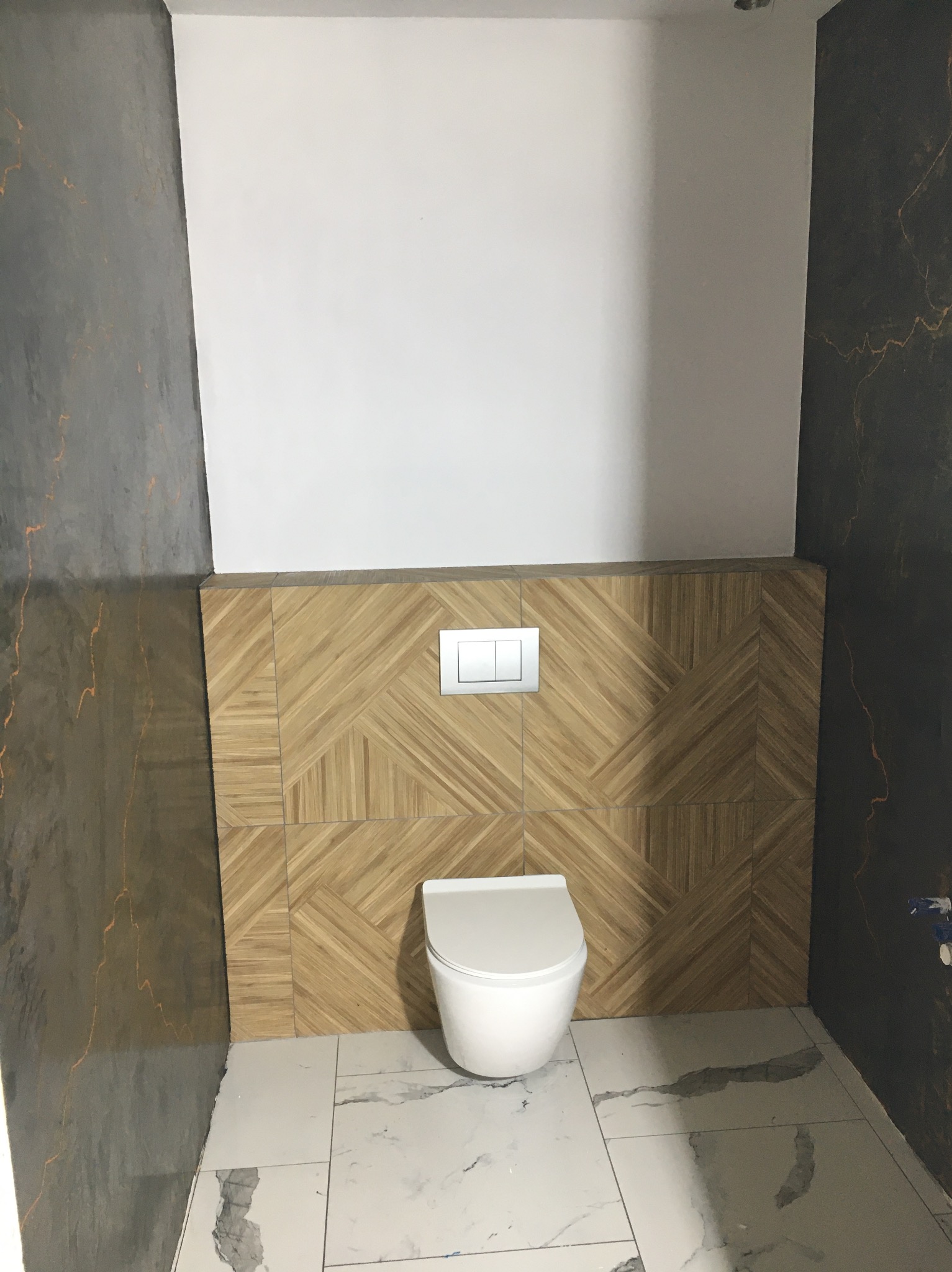 Nowoczesna toaleta podwieszana z drewnopodobną mozaiką na ścianie i marmurową posadzką w odcieniach szarości i bieli, z kontrastującymi ścianami.