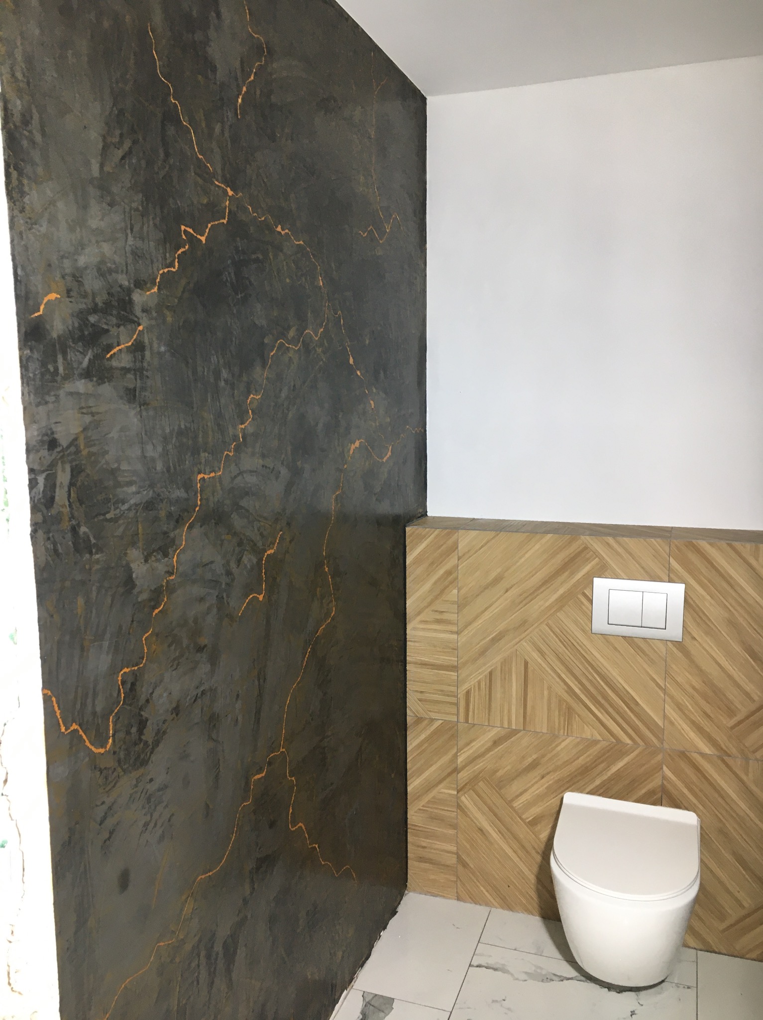 Wykończona ściana w łazience z dekoracyjnym tynkiem w odcieniach szarości i złotymi żyłkami, połączona z drewnopodobnymi płytkami i białą ceramiką.