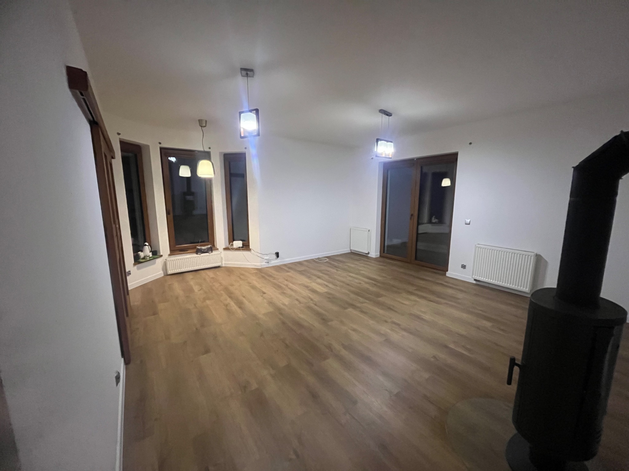 Jasne wnętrze po remoncie: drewniana podłoga, białe ściany, okna z drewnianymi ramami, nowoczesne lampy i piec wolnostojący. Minimalistyczny design.