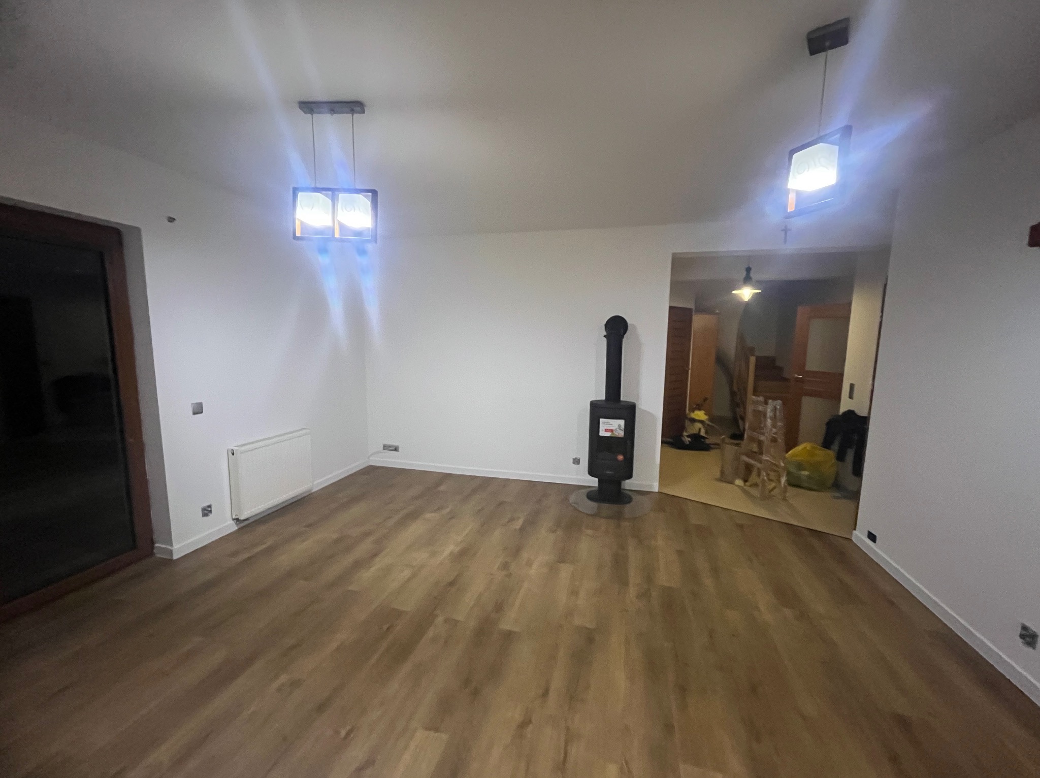 Jasne wnętrze po remoncie: drewnopodobna podłoga, białe ściany, nowoczesny piec wolnostojący i designerskie lampy wiszące. Widok na sąsiednie pomieszczenie.
