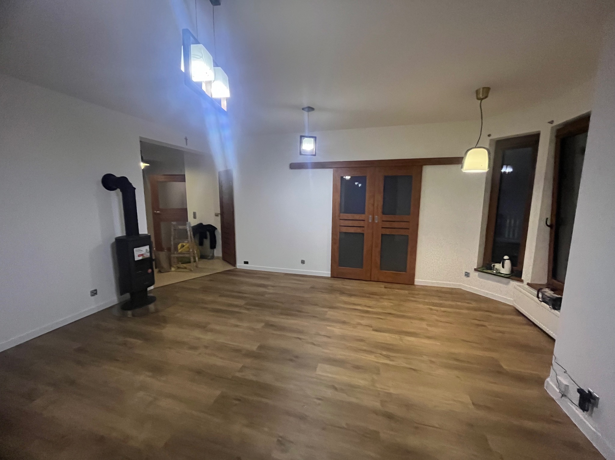 Jasne wnętrze z drewnianą podłogą, piecem wolnostojącym i przesuwnymi drzwiami. Widoczne okna z dekoracjami i nowoczesne oświetlenie sufitowe.