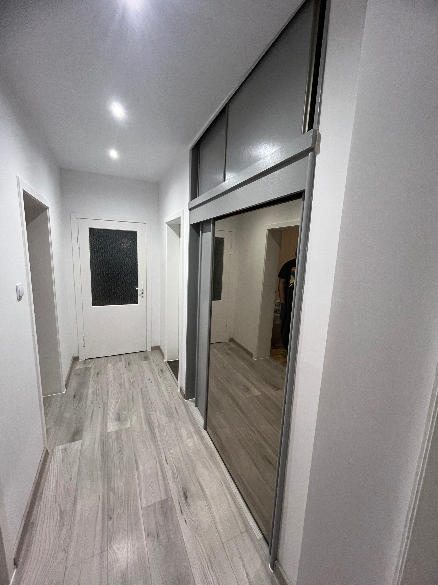 Jasny korytarz po remoncie z szarą podłogą imitującą drewno i szafą wnękową z lustrzanymi drzwiami przesuwnymi. Widoczne drzwi wejściowe i oświetlenie sufitowe.