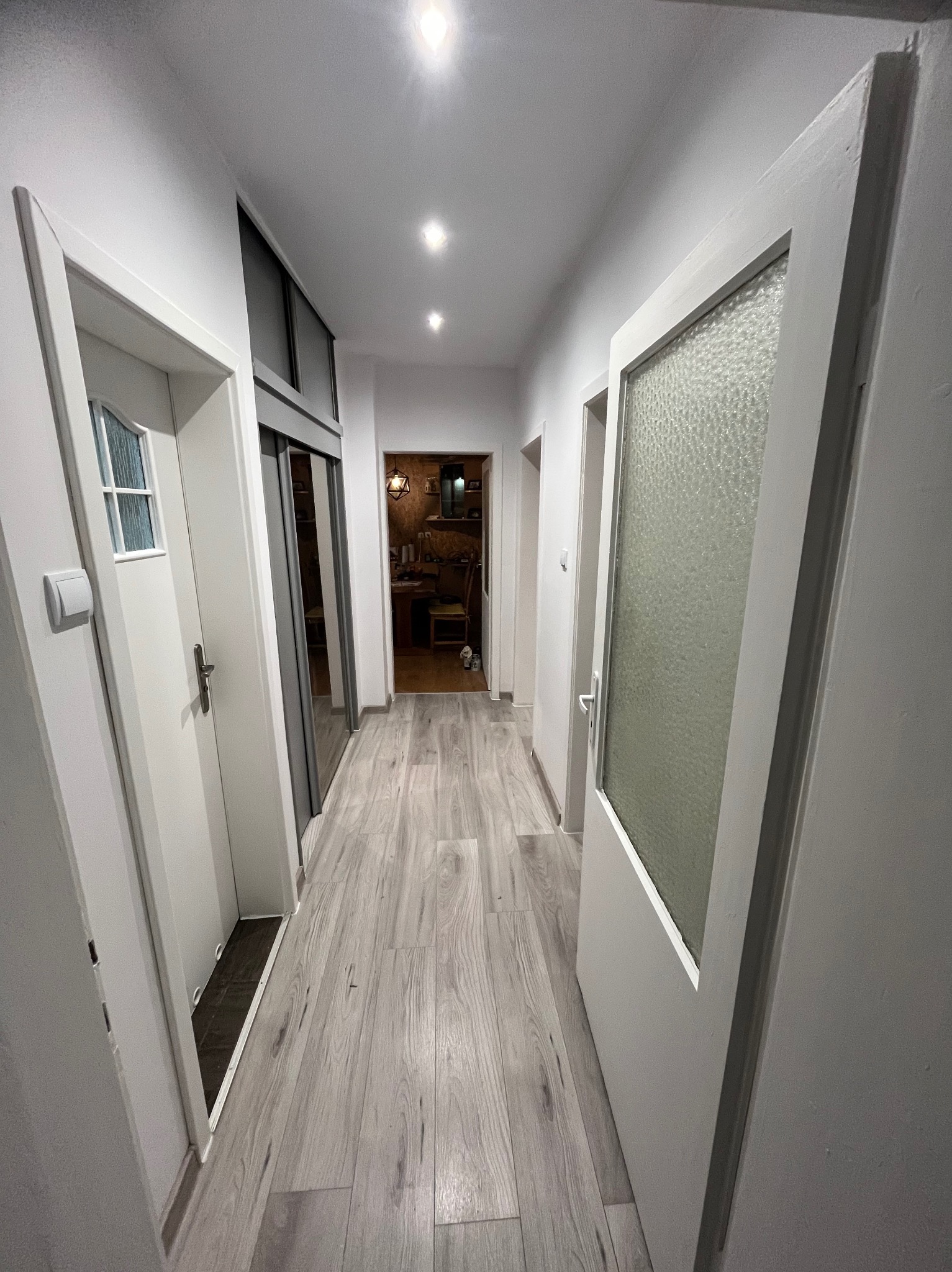 Jasny korytarz po remoncie z panelami podłogowymi imitującymi drewno, białymi ścianami i otwartymi drzwiami z przeszkleniami. Widok w głąb mieszkania.