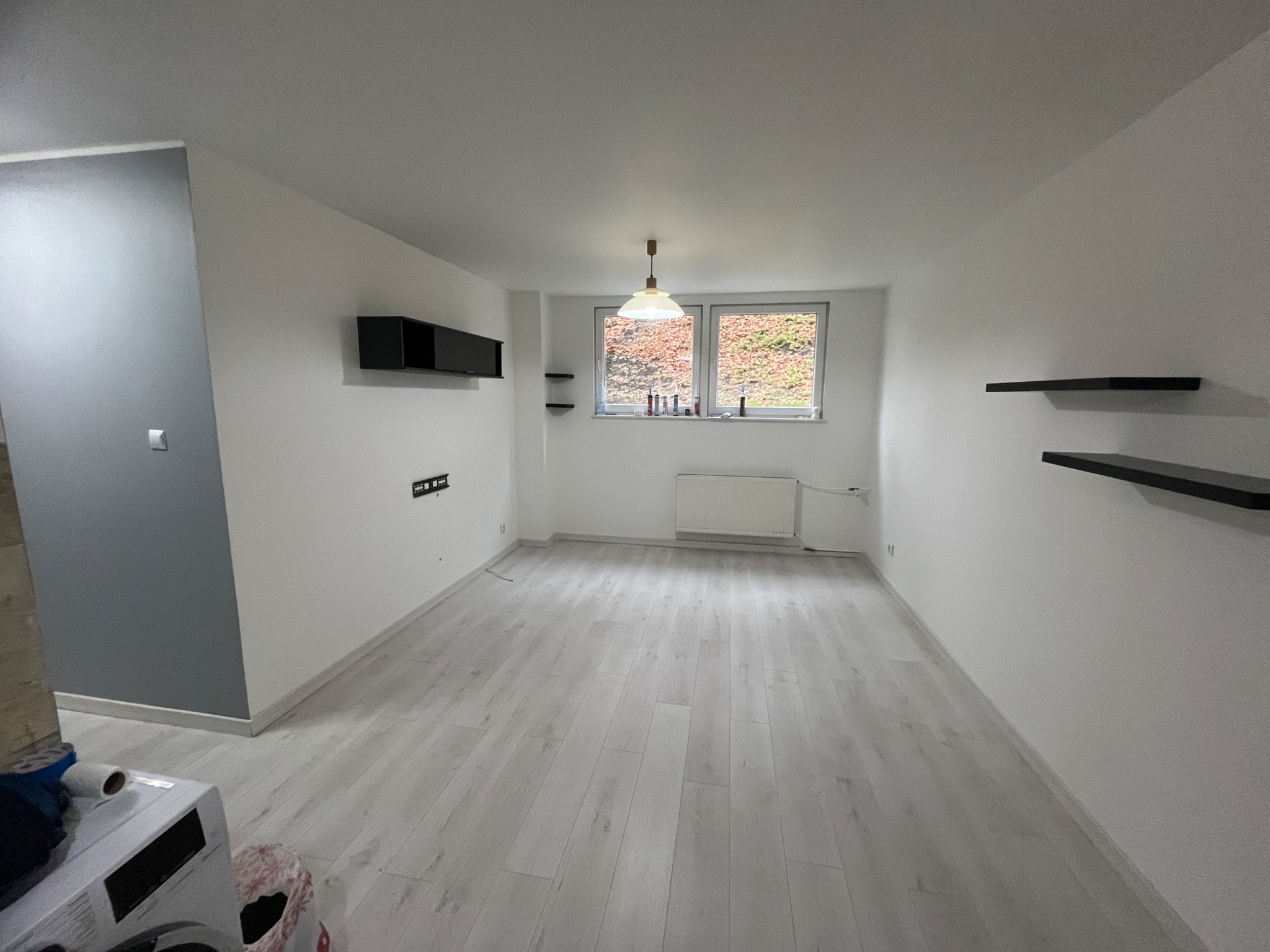 Jasne, przestronne wnętrze po remoncie: białe ściany, panele podłogowe, okna z widokiem na zieleń, czarne półki i szafka. Minimalistyczny design, lampa sufitowa.