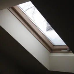 Zamontowane okno dachowe z drewnianą ramą, widok na zewnątrz w pochmurny dzień, fragment ściany wykończonej płytą karton gips.