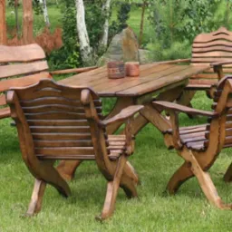 Zestaw rustykalnych mebli ogrodowych z litego drewna na trawniku, składający się z ławki, stołu i czterech krzeseł o charakterystycznych, falistych oparciach i krzyżowych nogach.