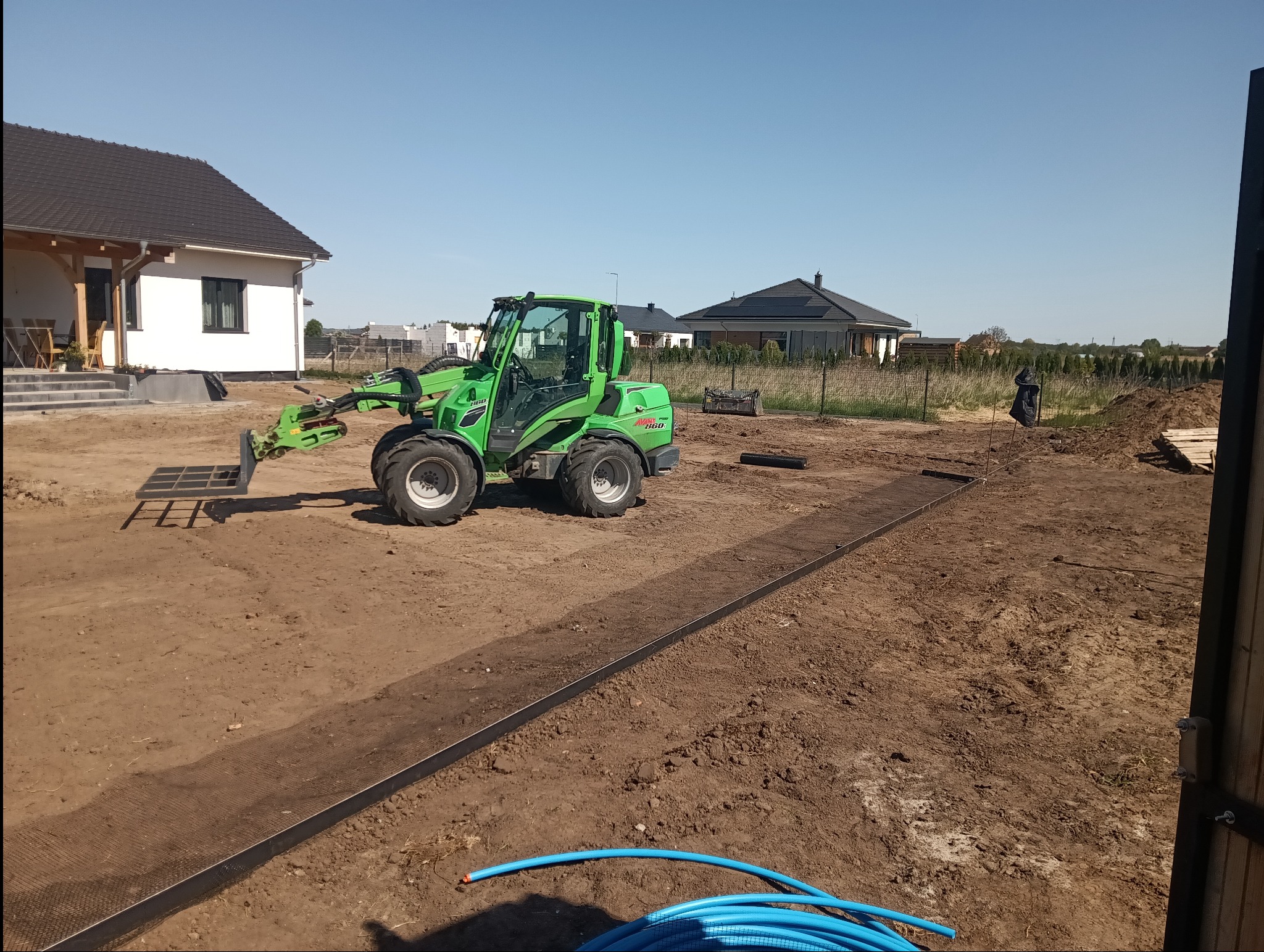 Teren przygotowany pod budowę: zielona ładowarka Avant 860 z osprzętem, obrzeża trawnikowe i zwoje niebieskich rur na ziemi, w tle domy jednorodzinne.