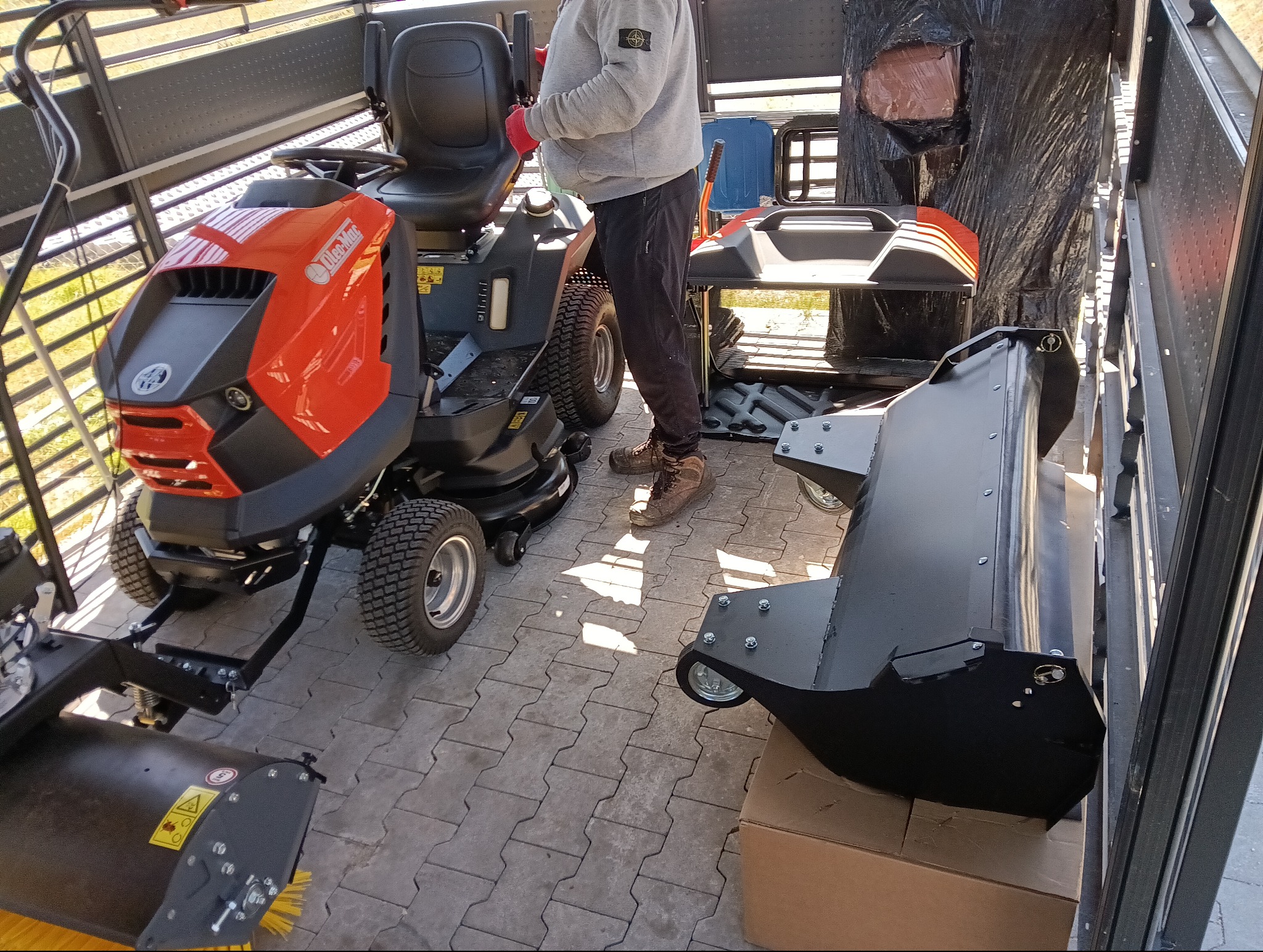 Pomarańczowy traktorek do trawy Oleo-Mac z osprzętem (pług, szczotka) w przestrzeni ładunkowej, widok z boku, kostka brukowa na podłożu.