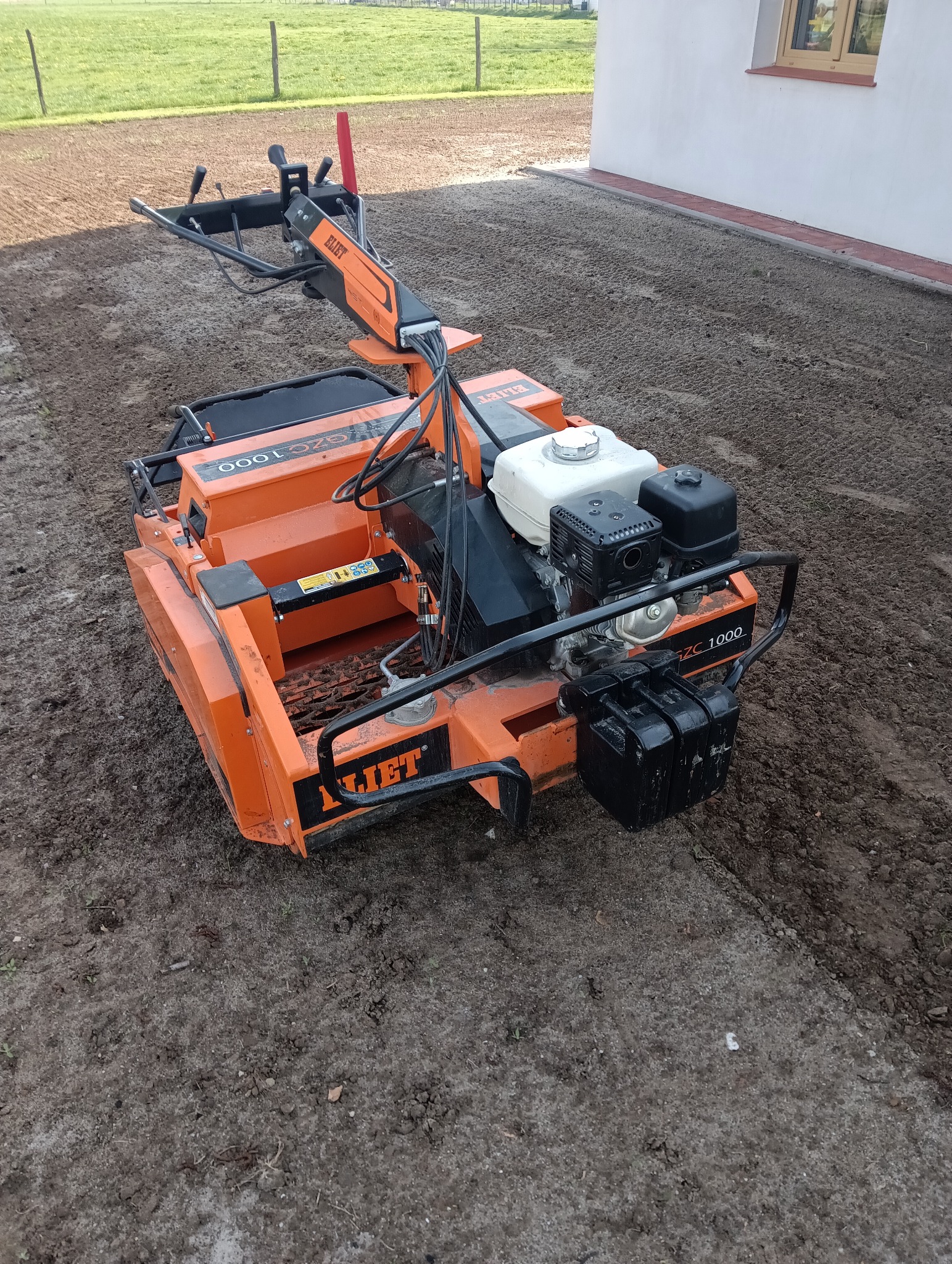 Pomarańczowa glebogryzarka marki Eliet na przygotowanym podłożu pod trawnik, obok budynku z oknem. Widoczne ślady po pracy maszyny.
