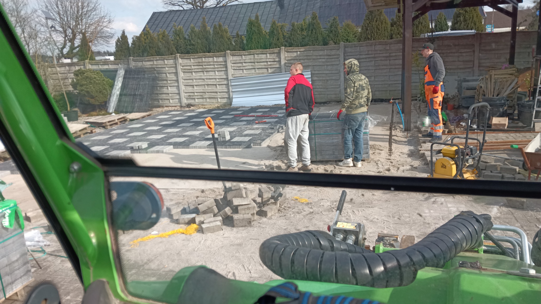 Układanie kostki brukowej na posesji. Widok z kabiny maszyny roboczej na pracowników i przygotowany teren. Częściowo ułożona kostka, palety z kostką, narzędzia.